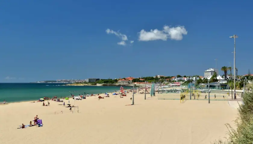 Amplia playa de arena dorada cerca de Lisboa, con gente disfrutando del sol y el mar. Hay una cancha de voleibol y edificios al fondo.