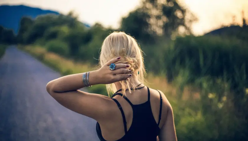 Blondynka z pierścionkiem w kształcie kwiatka, jak mały szelma, poprawia włosy na tle drogi i zieleni.