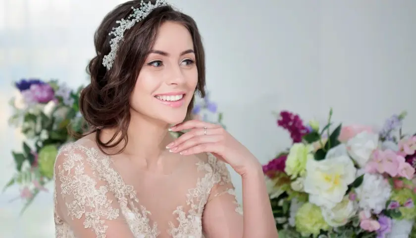Uśmiechnięta panna młoda z elegancką fryzurą ślubną z opaską, w koronkowej sukni, otoczona bukietami kwiatów.