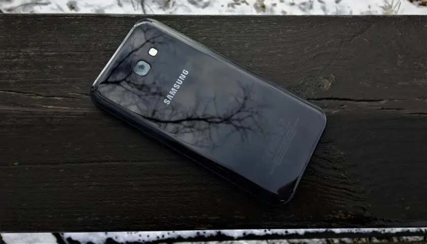 Czarny Samsung SM-A520F leży na drewnianej ławce, odbijając gałęzie drzew.