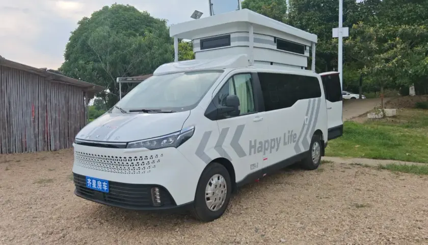Ce camping-car 9m blanc, avec "Happy Life" écrit dessus, est prêt pour l'aventure. Il est garé sur un chemin de gravier, à côté d'une structure en bois.
