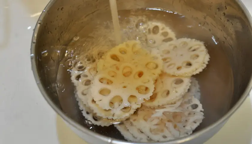 Slices of pickled lotus root are being rinsed in a metal bowl under running water, ready for a delicious meal.
