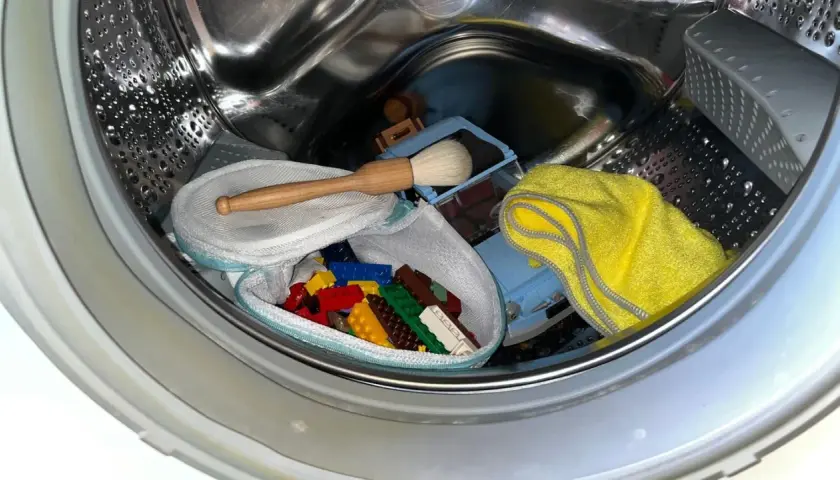 Bunte Duplo-Steine in einem Wäschenetz, ein Spielzeugauto, eine Bürste und ein gelbes Tuch warten auf die Reinigung in der Waschmaschine.