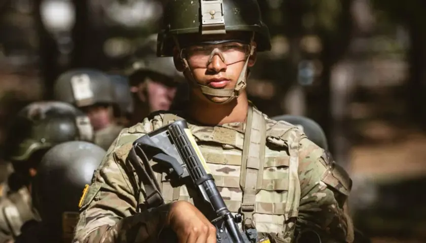 A young soldier, determined and focused, stands ready during far bootcamp. He wears a helmet, protective eyewear, and camouflage fatigues, holding his rifle.