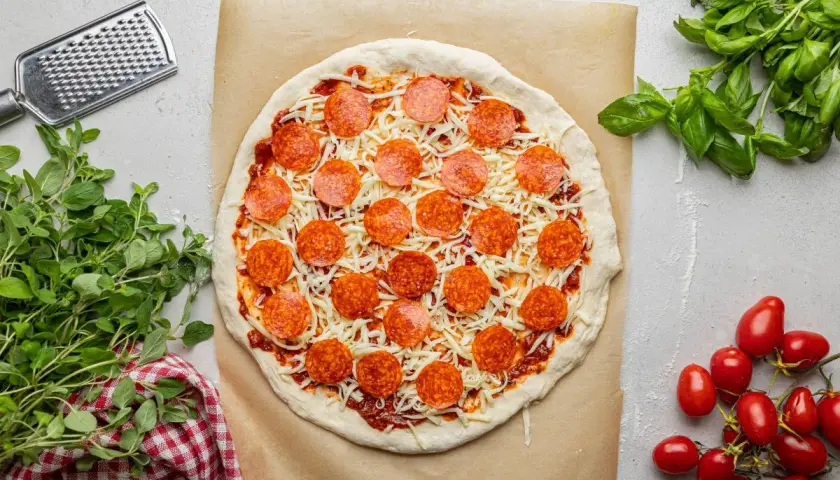 Pizza z sosem, serem i okrągłymi plasterkami pepperoni, gotowa do pieczenia. Obok świeża bazylia i pomidorki.