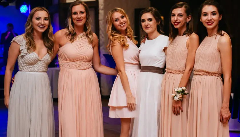 Uśmiechnięte druhenki w pastelowych sukniach pozują do zdjęcia. Jedna z nich ma na ręce wianek z kwiatów.
