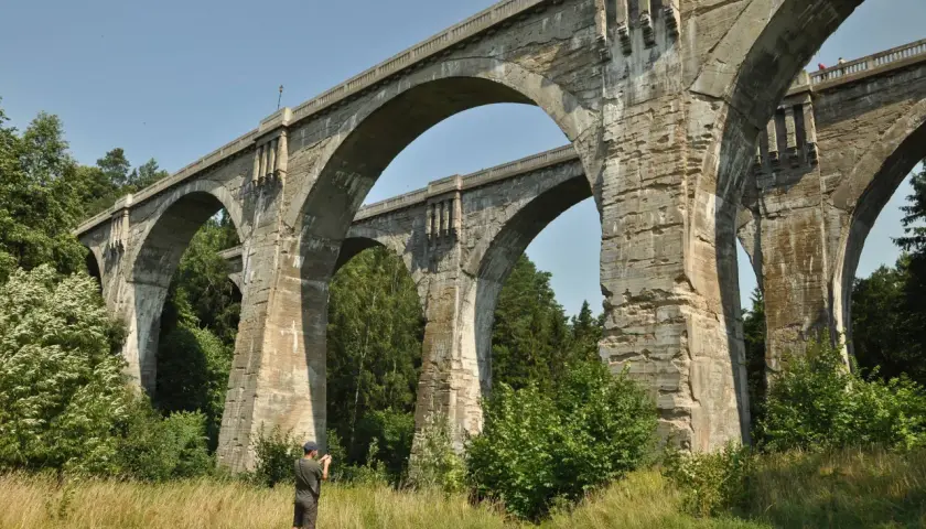 Imponujące mosty w Stańczykach, zbudowane z kamienia, górują nad zielonym krajobrazem. Mężczyzna z aparatem dokumentuje ich monumentalność.
