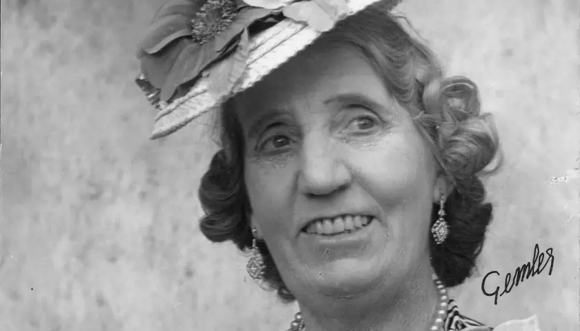 Julia Cæsar, en glad dam med hatt och örhängen, ler mot betraktaren.