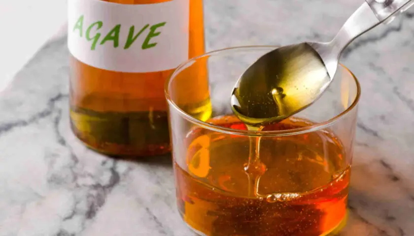 Cuchara con sirope de agave cayendo en un vaso, junto a una botella etiquetada.