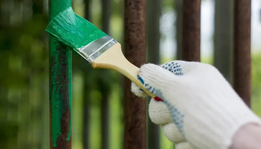 Mano guantata stende vernice verde su un palo di ferro arrugginito. La temperatura ideale per verniciare il ferro è fondamentale per una buona resa.