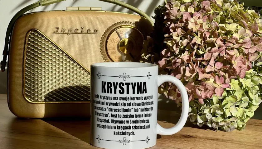 Biały kubek z napisem "Krystyna" na tle starego radia i bukietu suszonych hortensji.