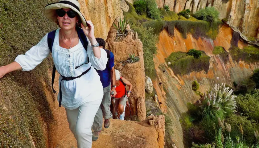 Hikers ascend a rocky path during hiking trips in Portugal, with lush green vegetation clinging to the orange cliffs.