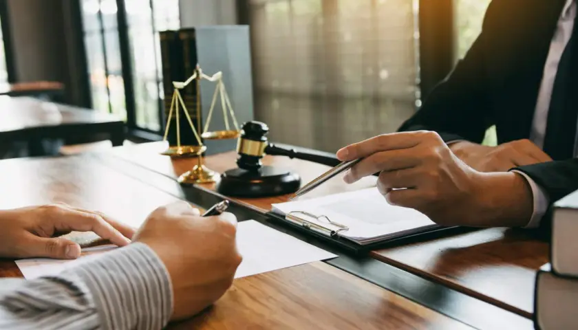 Dwóch mężczyzn przy biurku, jeden podpisuje dokument, drugi wskazuje piórem. W tle waga sprawiedliwości i młotek. Po co notariusz przy wynajmie mieszkania? By mieć pewność prawną.