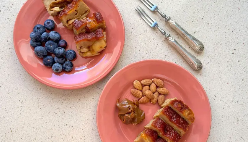 Dos platos rosados con porciones de pudin de manzana, arándanos y almendras. Un postre casero para disfrutar.
