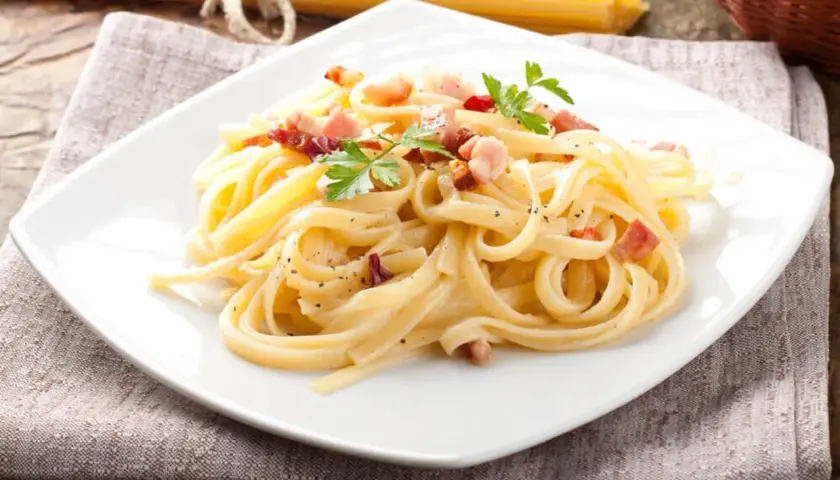 Pyszne spaghetti carbonara z boczkiem i natką pietruszki na białym talerzu, obok surowy makaron.