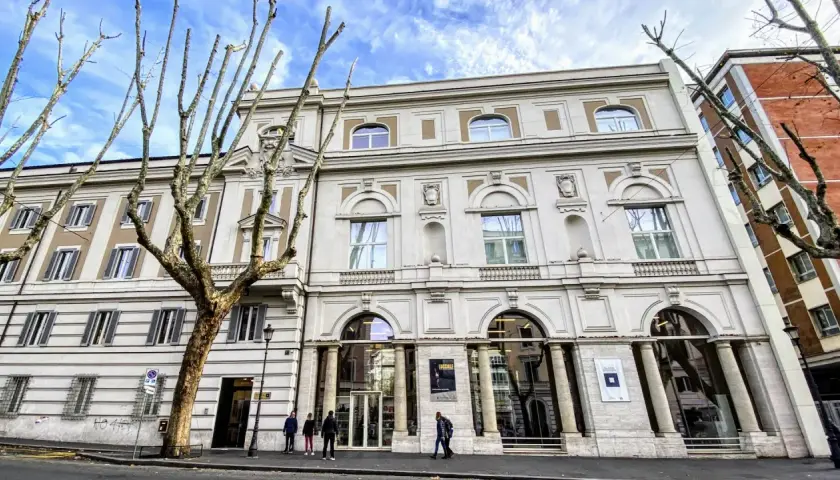 Le **Palazzo Merulana**, un édifice élégant, se dresse sous un ciel bleu nuageux, orné d'arbres dénudés aux branches graphiques.