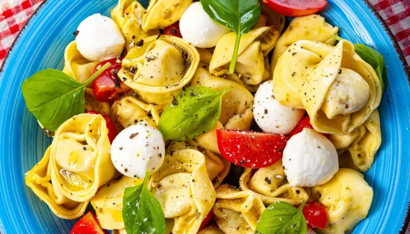 Ein bunter Tortellini Salat mit Mozzarella-Kugeln, Tomatenstücken und frischem Basilikum, appetitlich in einer blauen Schüssel angerichtet.