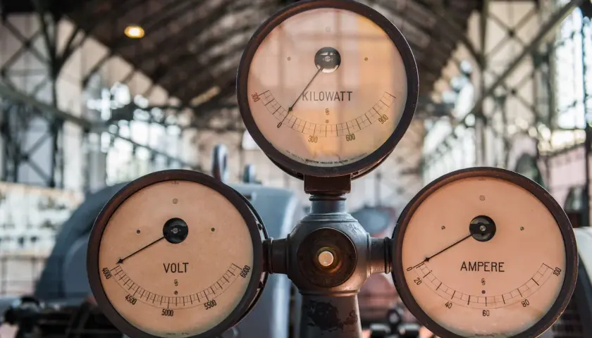 Trzy zabytkowe mierniki: VOLT, AMPERE i KILOWATT, wskazujące wartości prądu i mocy.