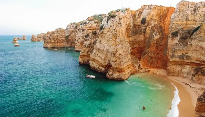 Aguas turquesas y acantilados dorados en las **playas Algarve**. Una pequeña barca navega cerca de una cala escondida donde una persona nada.