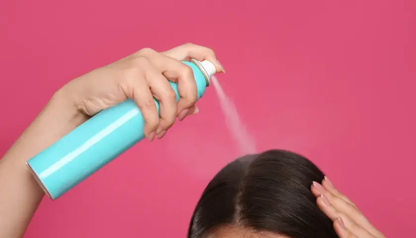 Dłoń trzymająca turkusowy spray z suchym szamponem, rozpylająca go na ciemne włosy.