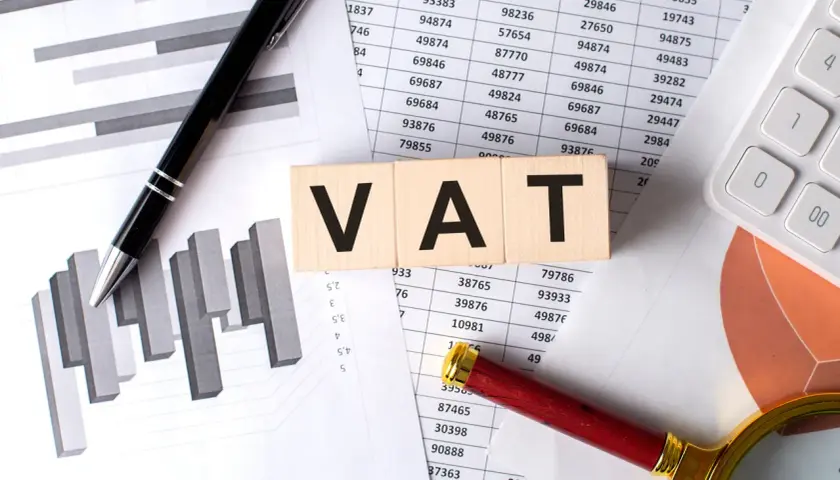 Drewniane klocki z napisem VAT leżą na dokumentach z wykresami i tabelami. Jak odliczyć VAT z faktury? Pióro i lupa sugerują analizę.