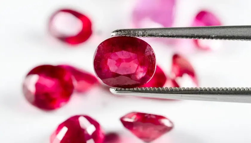 Ein Juwelier hält mit einer Pinzette einen tiefroten Edelstein, der wie der seltenste Diamant der Welt aussieht. Mehrere andere rote Steine liegen unscharf im Hintergrund.