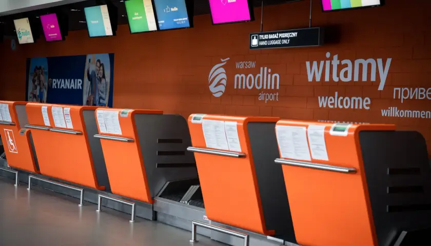 Pomarańczowe stanowiska odprawy bagażowej na lotnisku Modlin z tablicami informacyjnymi i logo lotniska.