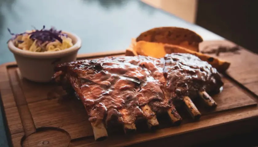 Délicieuses côtes levées au bbq, nappées de sauce, servies avec une salade de chou et des quartiers de pommes de terre sur une planche en bois.