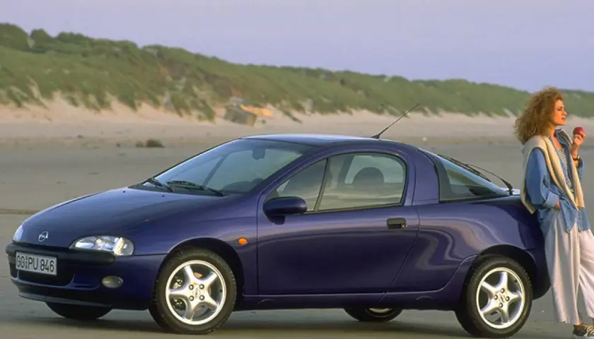 Una donna appoggiata a una Opel Tigra blu, icona delle macchine anni 90, sulla spiaggia al tramonto.
