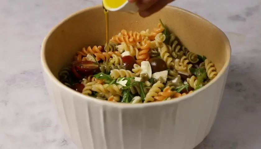 Un bol lleno de pasta de colores, tomates cherry, queso y rúcula, con aceite de oliva vertiéndose sobre ella.