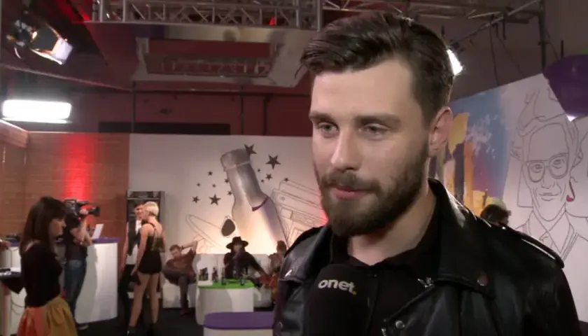Ernest Staniaszek udziela wywiadu na tle scenografii z butelką i gwiazdami.