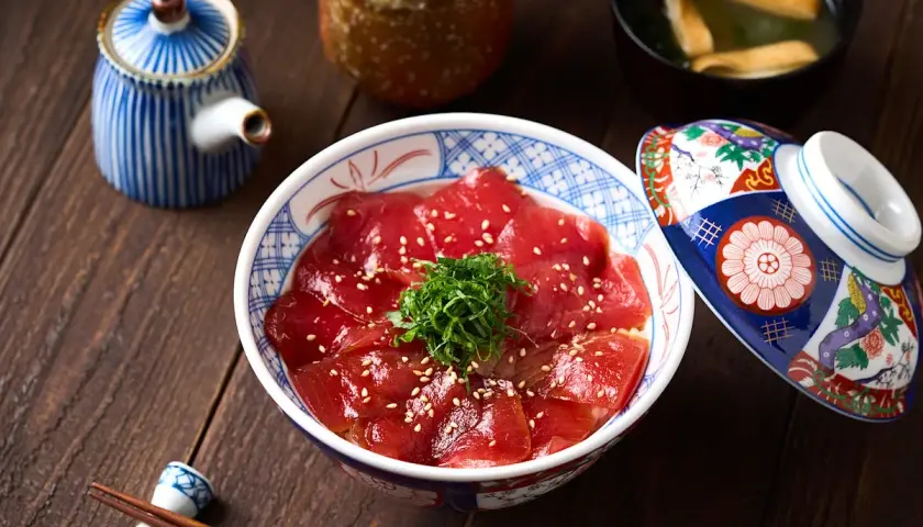 A bowl of delicious Japanese sweet rice topped with thinly sliced tuna, sesame seeds, and green garnish. A decorative lid rests beside it.