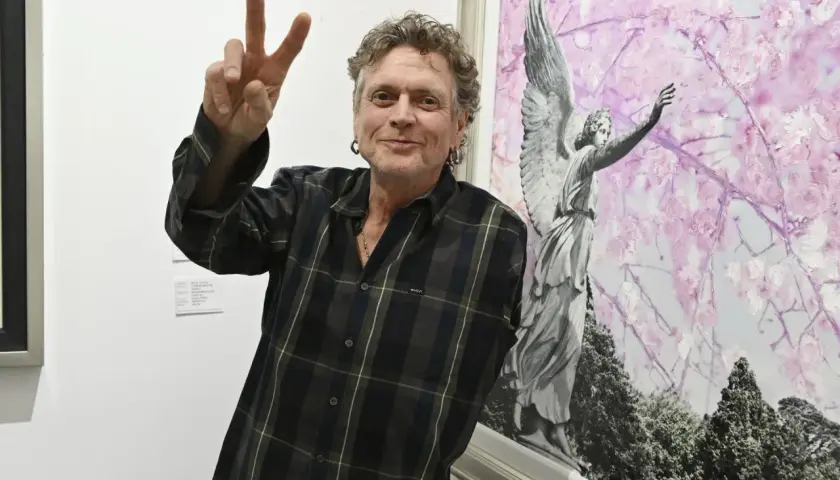 Rick Allen, perkusista Def Leppard, pozuje z uniesionym znakiem pokoju obok obrazu z aniołem i kwitnącymi drzewami.