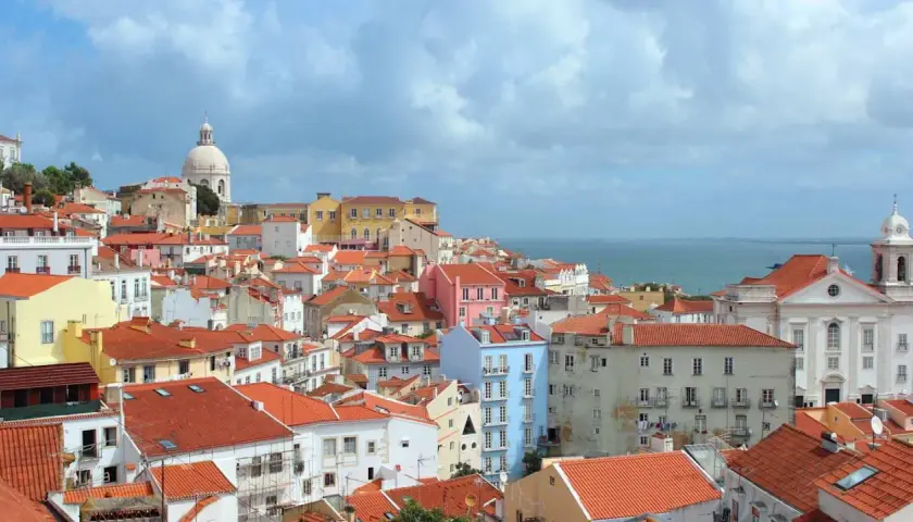 One day in Lisbon, a panorama of colorful buildings with red-tiled roofs stretches towards the shimmering blue sea under a cloudy sky.