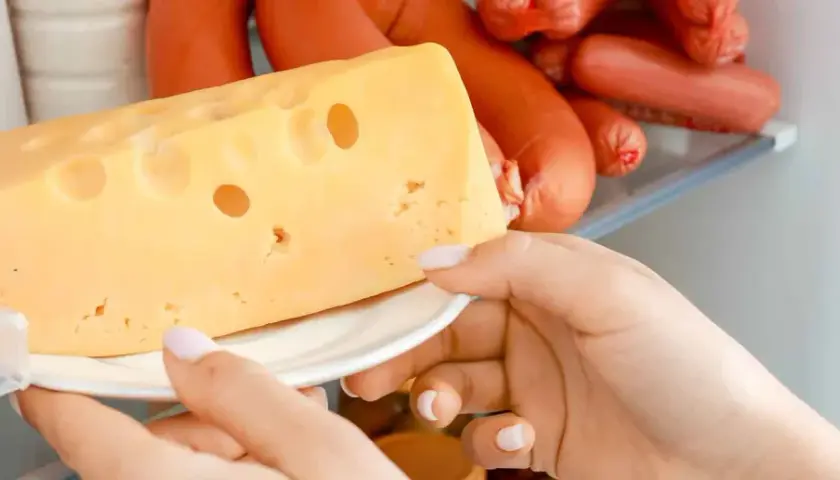 Mano prende un pezzo di formaggio svizzero da un frigorifero pieno di salumi. Formaggi salutari per uno spuntino.