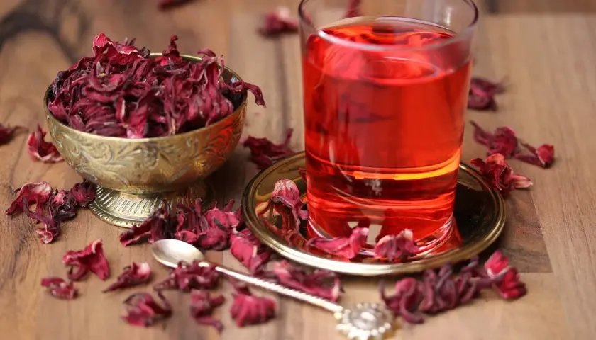 Ein Glas leuchtend roter Malva Sylvestris Tee steht neben einer Schale getrockneter Blüten. Getrocknete Blütenblätter sind auf dem Holztisch verstreut.