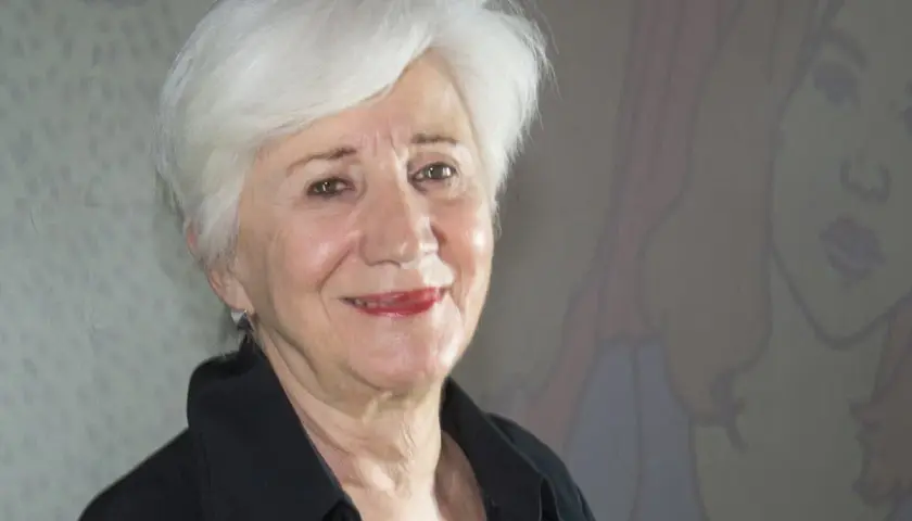 Olympia Dukakis ler vänligt mot kameran, med sitt karakteristiska vita hår och en svart skjorta. Bakom henne anas en tecknad figur.