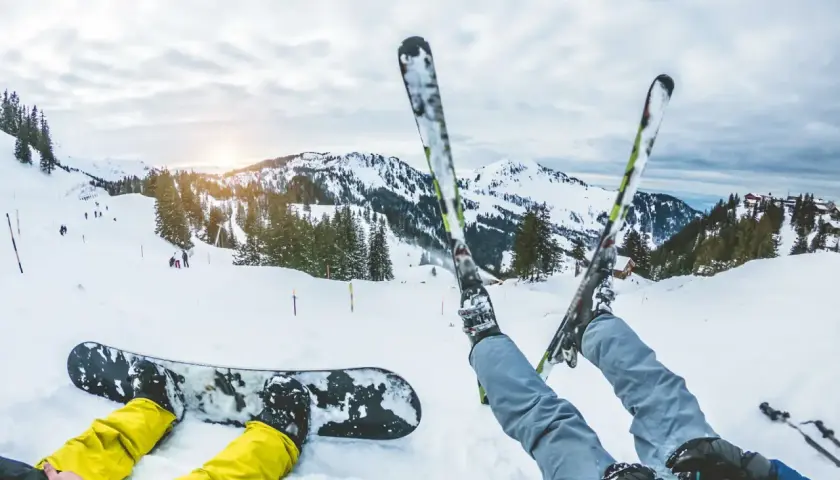 Dwie osoby odpoczywają na śniegu. Jedna leży na desce snowboardowej, druga ma założone narty. W tle góry i stoki narciarskie.