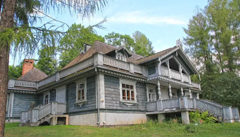 Drewniany dworek w parku pałacowym w Białowieży, z werandami i ozdobnymi detalami, otoczony zielenią.