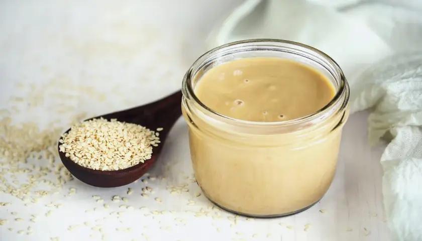 Un vasetto di tahina, crema di sesamo, accanto a un cucchiaio di semi. Scopri cos'è la tahina e come usarla.