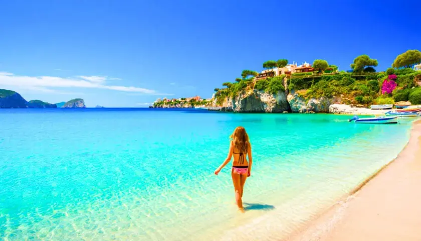 Femme en bikini entrant dans l'eau turquoise d'une plage idyllique, avec des maisons perchées sur une falaise, typique de la côte méditerranée en Italie.