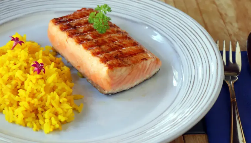 Un delicioso salmón a la plancha con marcas de parrilla, servido con arroz amarillo y decorado con perejil y flores moradas.
