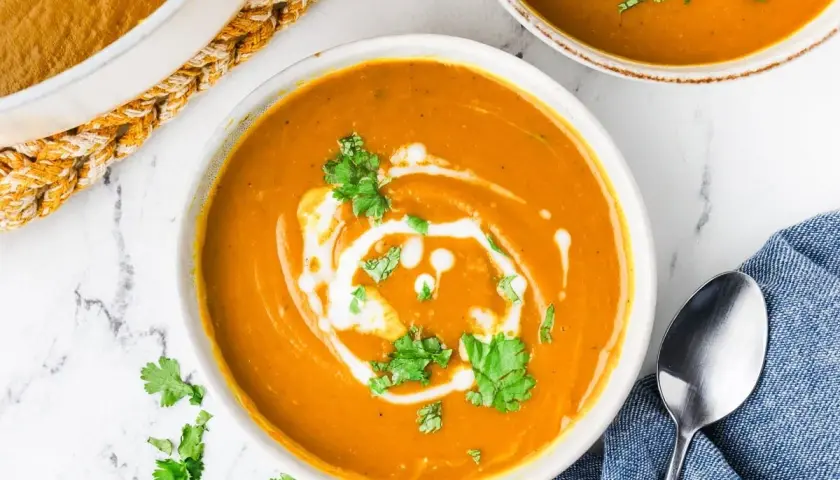 A creamy, vibrant bowl of kabocha squash soup, swirled with coconut cream and garnished with fresh cilantro. A spoon rests on a blue napkin beside it.