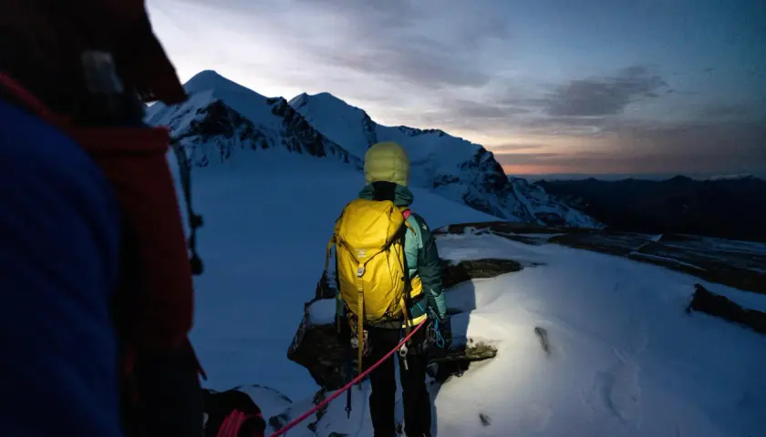 Alpinista z żółtym plecakiem lawinowym, gotowy na wyprawę w góry o wschodzie słońca. Plecak lawinowy jak działa? Zapewnia bezpieczeństwo w górach.