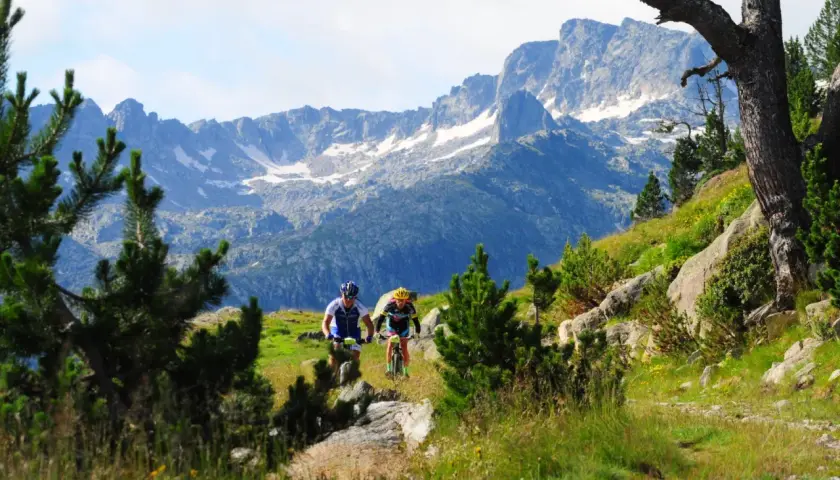 Ciclistas descienden por un sendero en el Pla de Beret, con imponentes montañas nevadas de fondo.