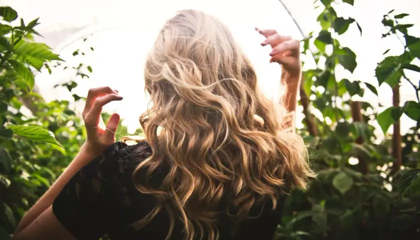 Blond włosy falują na wietrze, jakby przechodziły anwen test na porowatość. Kobieta w czarnej koronkowej bluzce stoi wśród zielonych liści.
