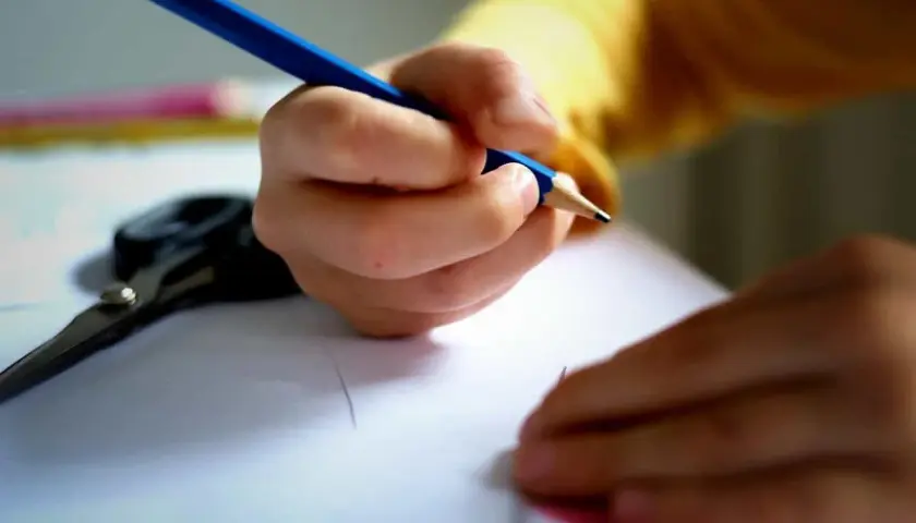 Dłoń podmiotu lirycznego trzyma niebieską kredkę, rysując na białej kartce. Obok leżą nożyczki.