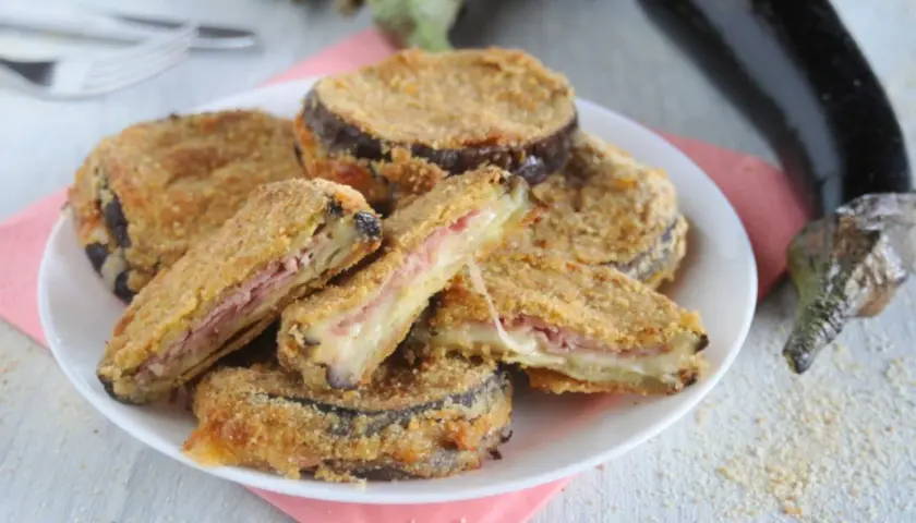 Cordon bleu di melanzane impanati e fritti, ripieni di prosciutto e formaggio filante, serviti su un piatto bianco.