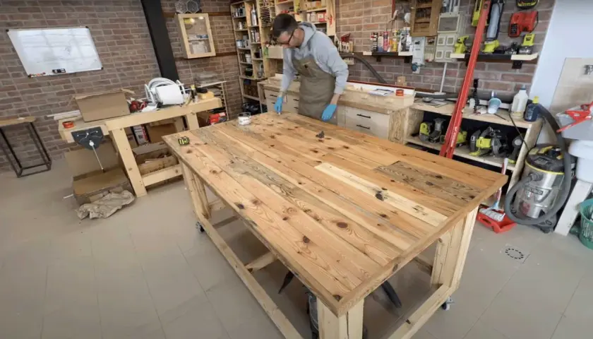 Uomo con guanti blu applica finitura su un robusto banco da lavoro in legno fai da te, circondato da attrezzi e materiali in un laboratorio.