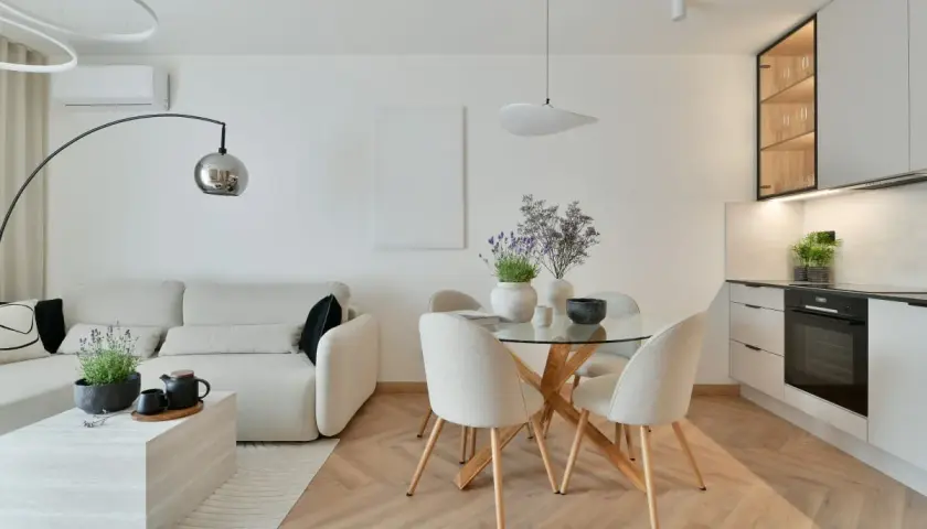 Nowoczesna aranżacja salonu z wygodną sofą, okrągłym stołem i aneksem kuchennym. Jasne kolory i drewniana podłoga tworzą przytulną atmosferę.